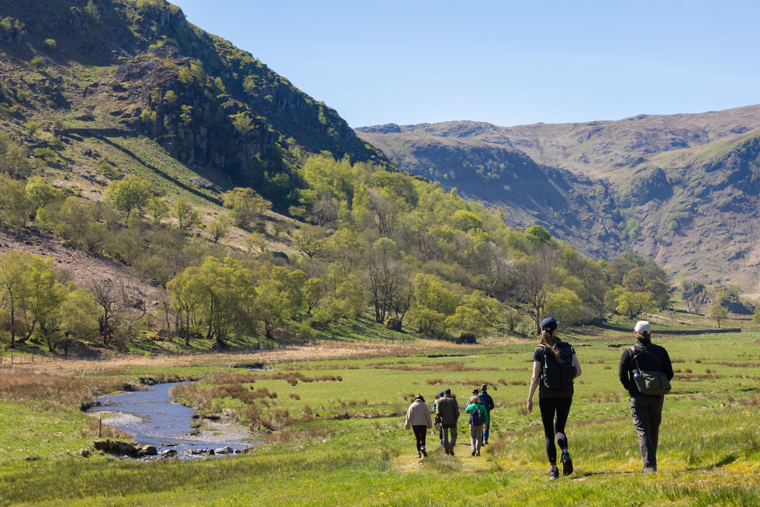 Photo of people walking across grassland besides a winding river with a back drop of hills.