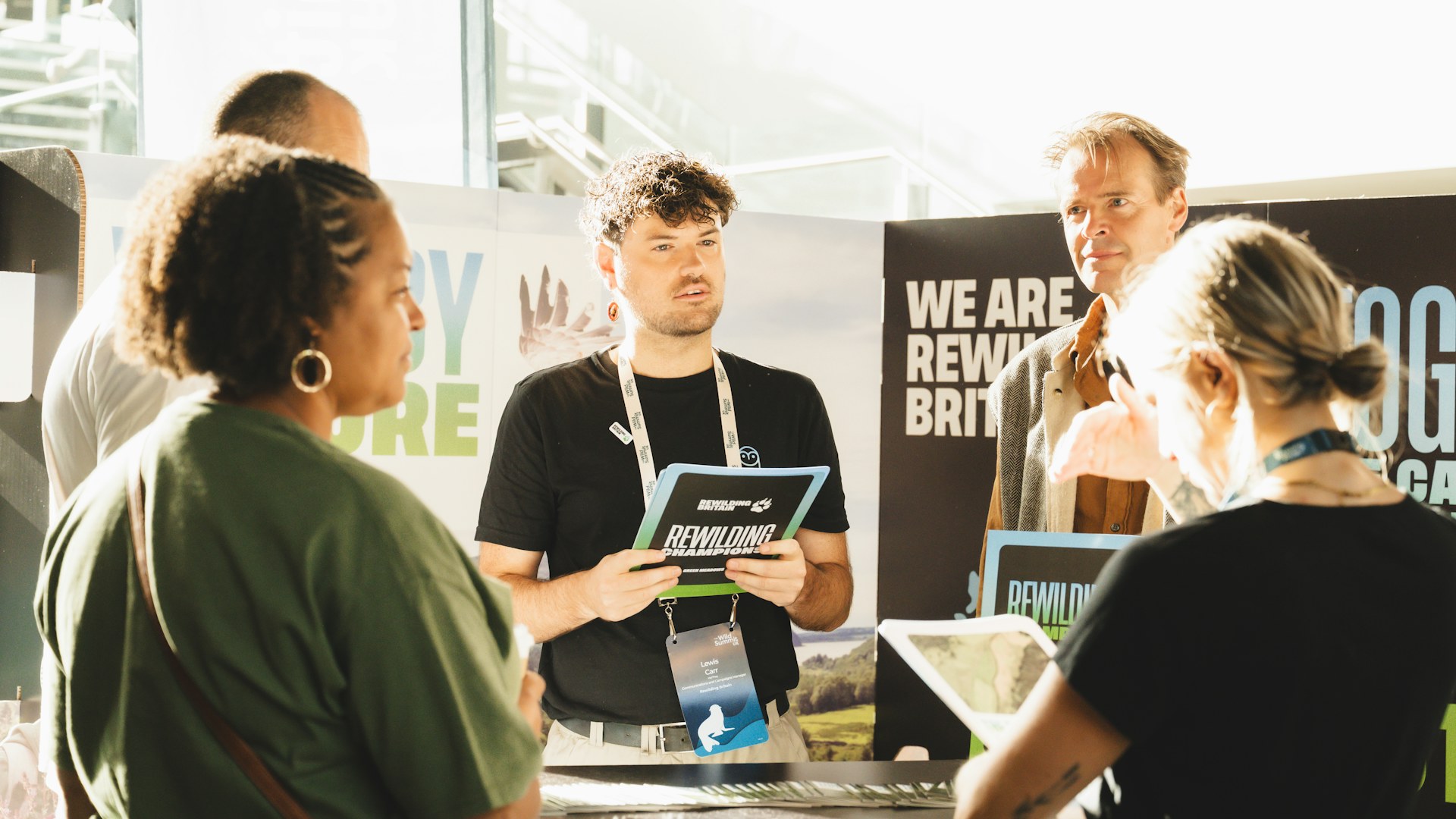 Rewilding Britain employee Lewis Carr stood in front of Rewilding Britain's stand at Wild Summit in Bristol talking to attendees.