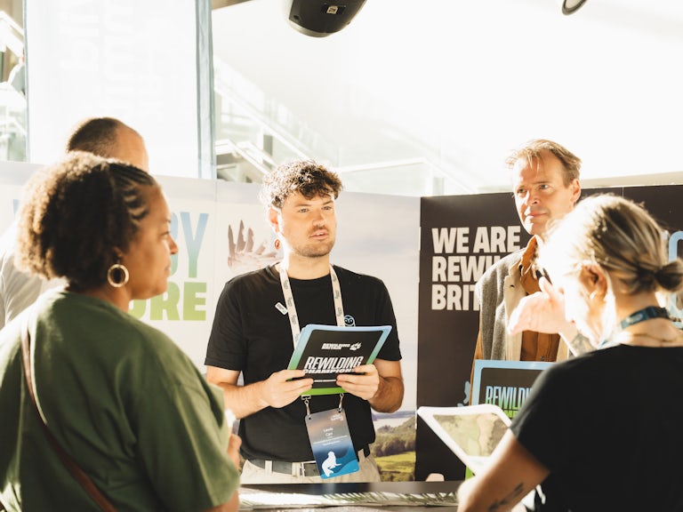Rewilding Britain employee Lewis Carr stood in front of Rewilding Britain's stand at Wild Summit in Bristol talking to attendees.