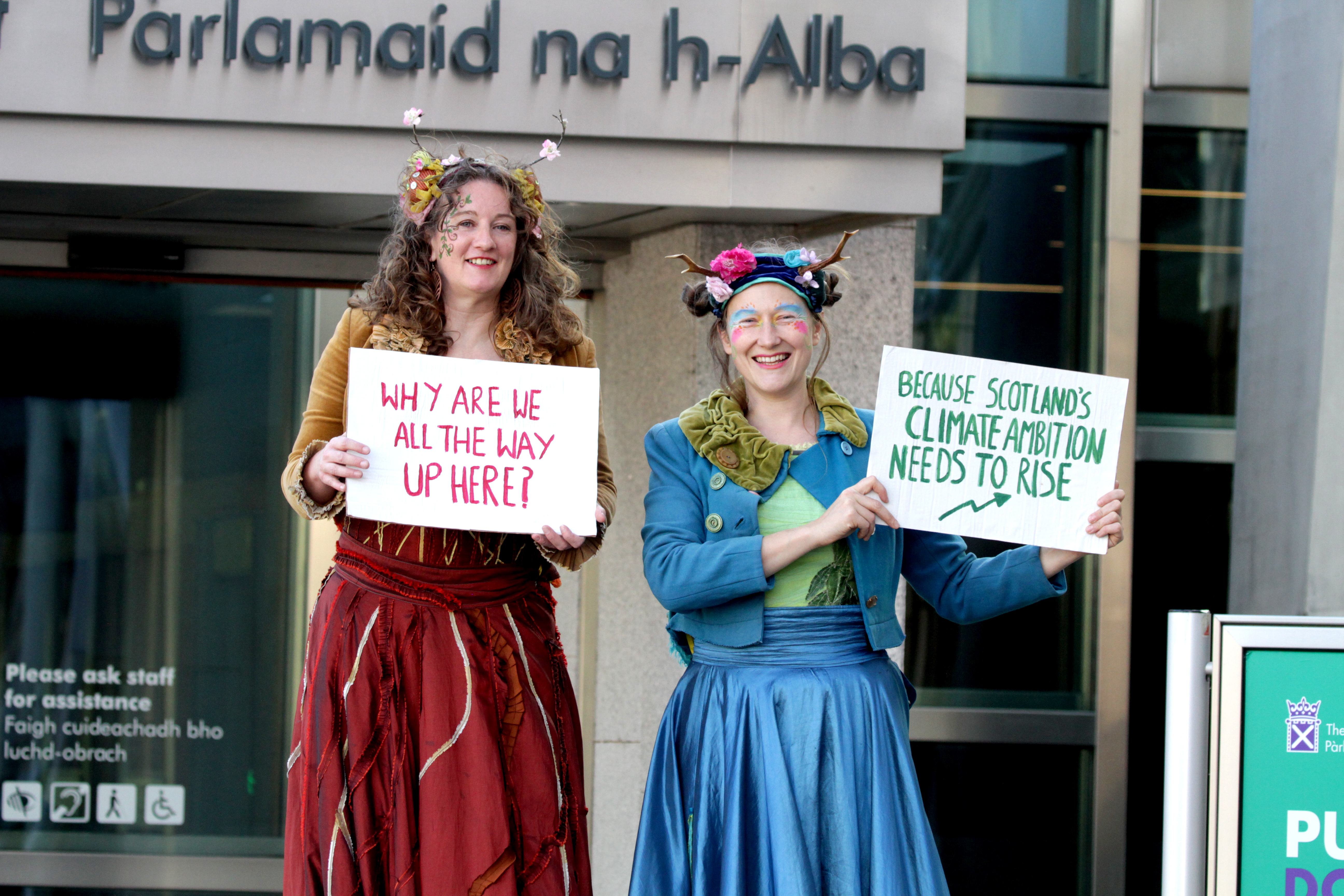 Two women stand holding protest signs on stilts outside Scottish Parliament