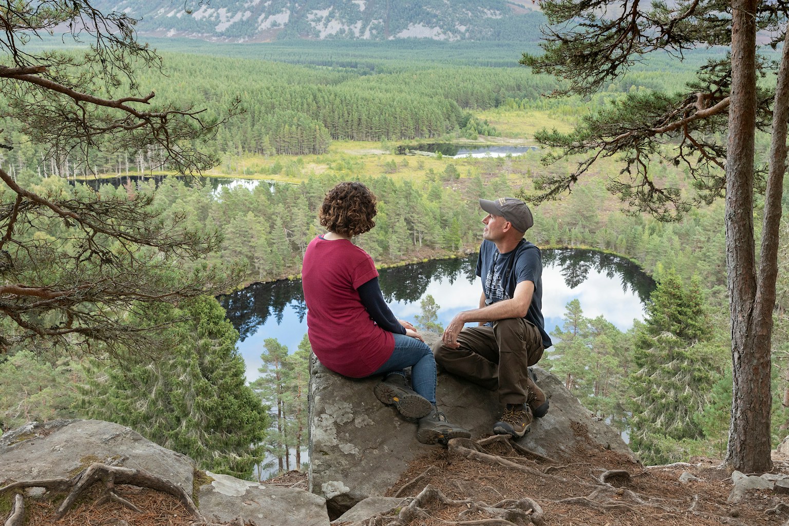 A man and a woman sat on a rock looking out from a scenic viewpoint above Uath Lochans in the Cairngorms National Park