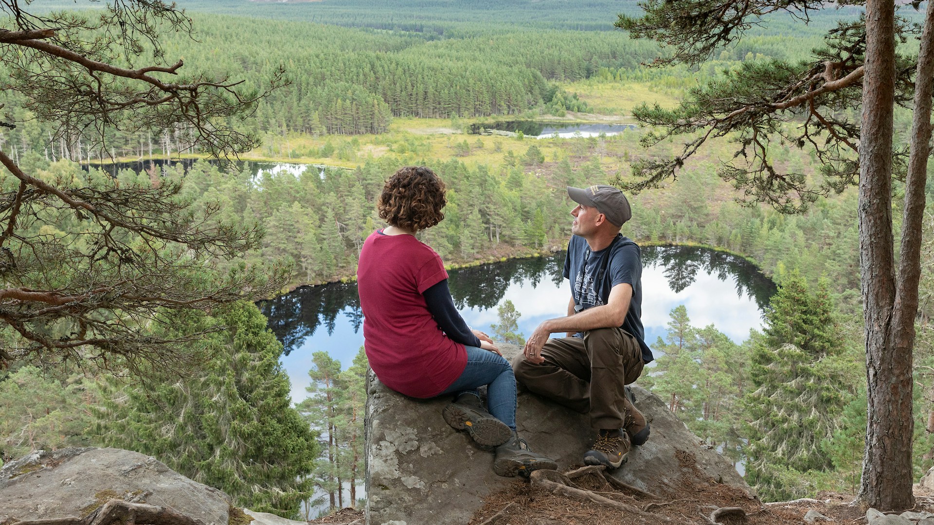A man and a woman sat on a rock looking out from a scenic viewpoint above Uath Lochans in the Cairngorms National Park