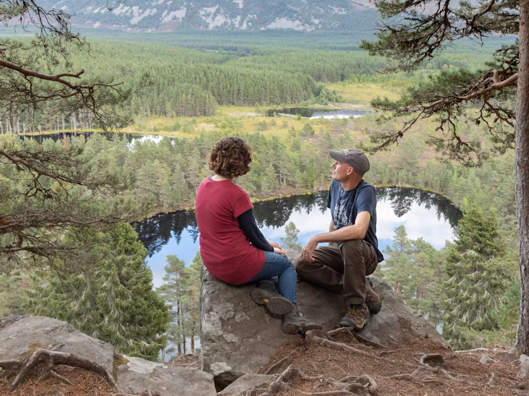 A man and a woman sat on a rock looking out from a scenic viewpoint above Uath Lochans in the Cairngorms National Park