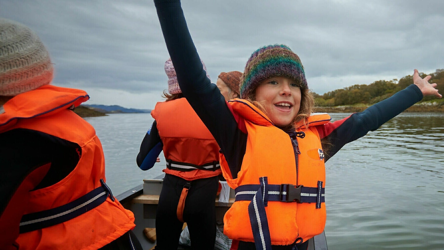 Child in orange life vest, hands up, in a boat with three other people volunteering for Seawilding