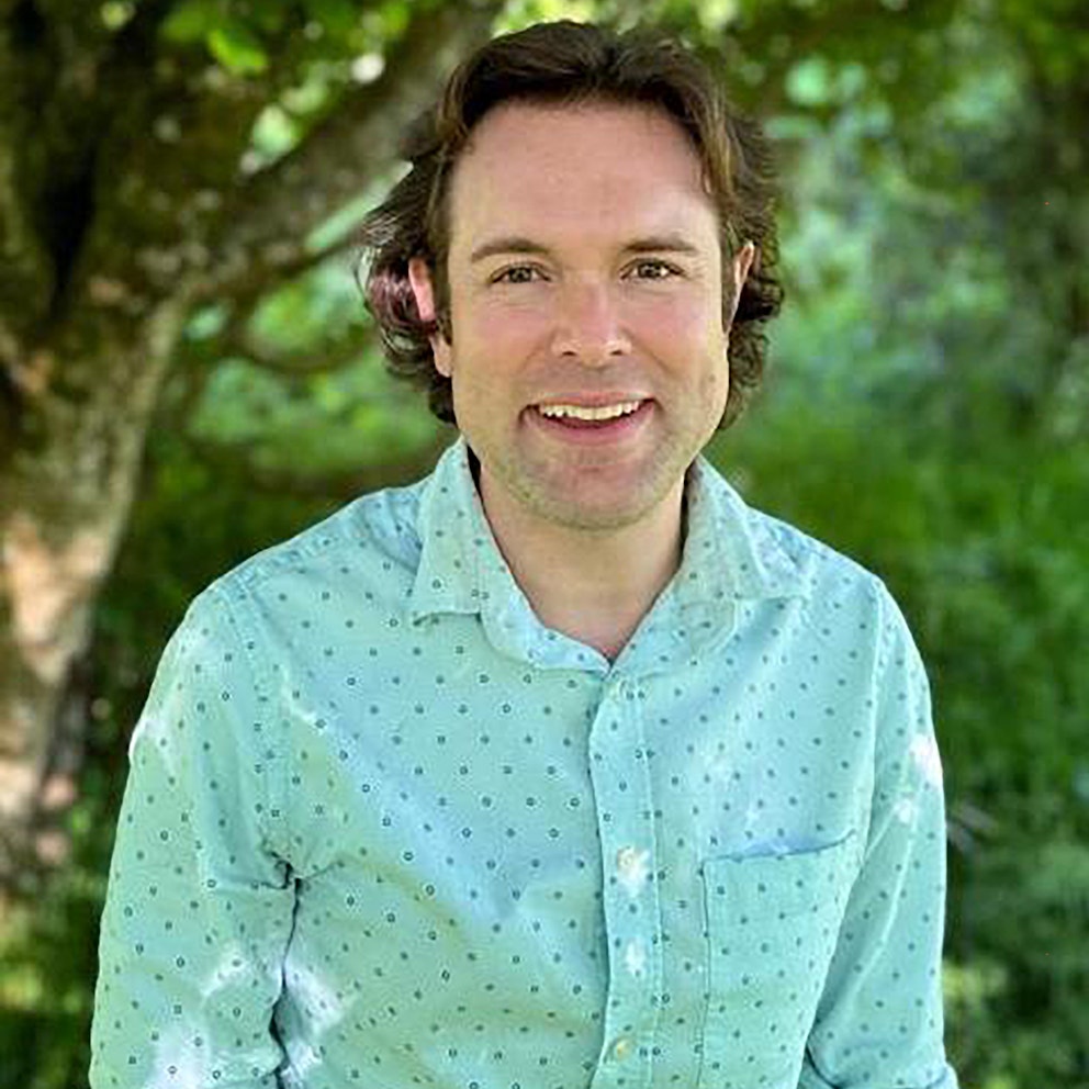 Close-up portrait of Ollie Newham wearing a light blue shirt standing in a woodland