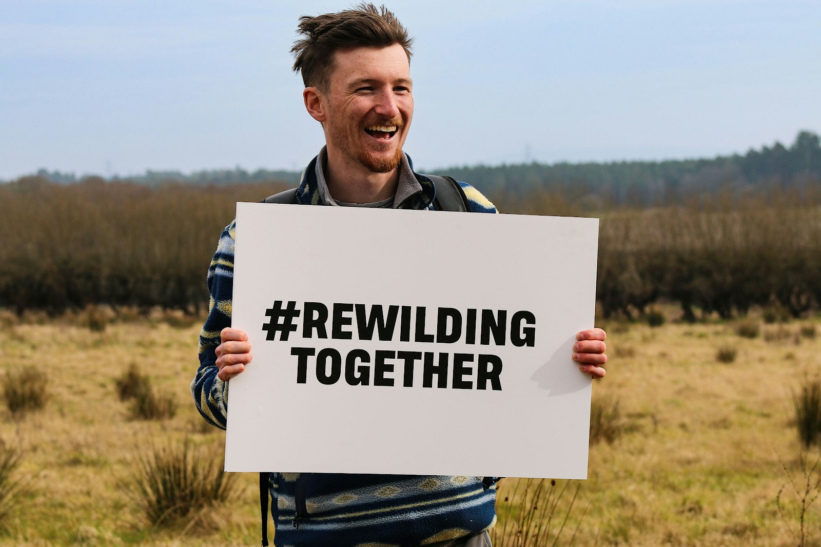 A man holding a sign saying #RewildingTogether in a field that's in rewilding